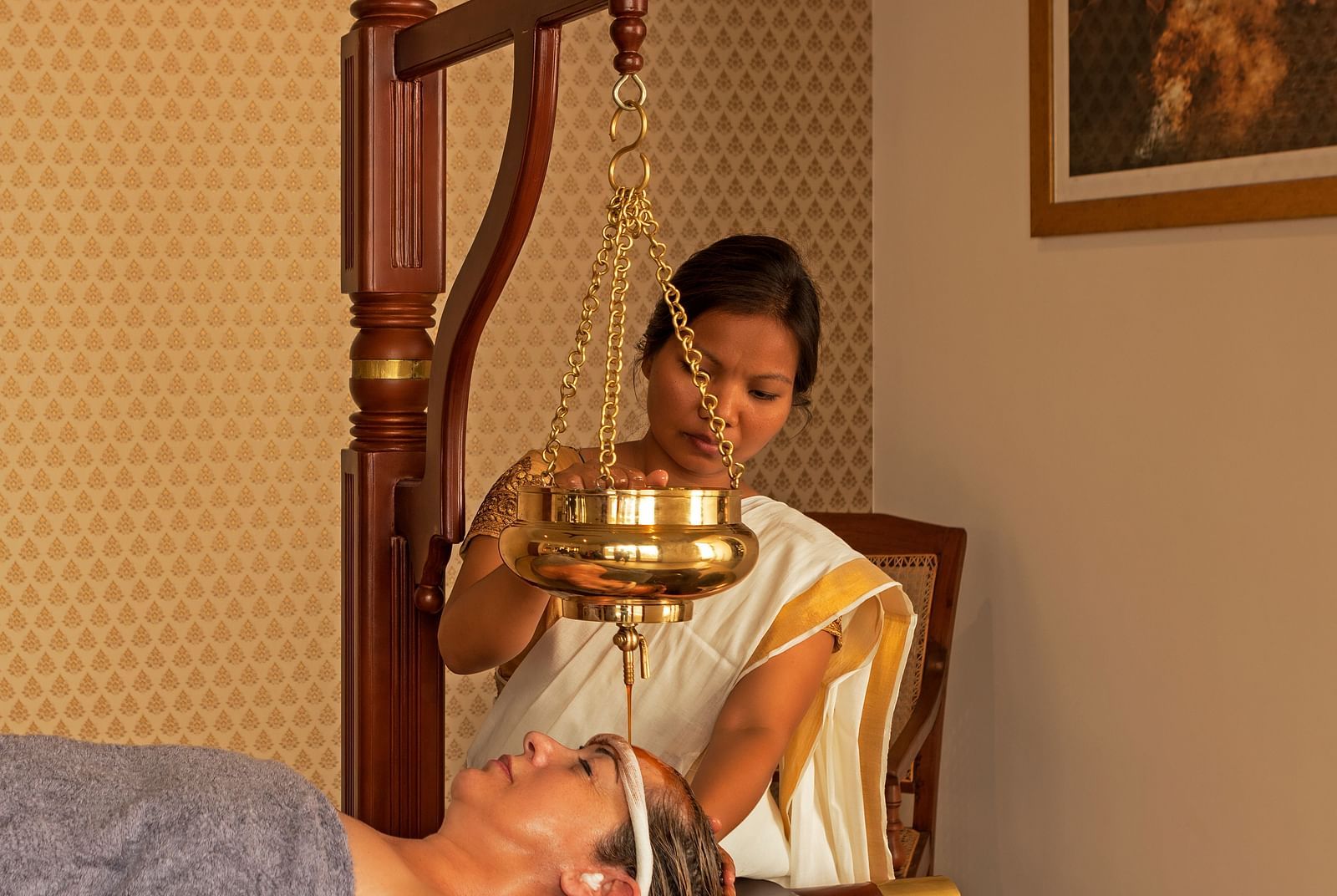 A person receiving a treatment in a room with a painting and wooden furniture - Shreyas Retreat