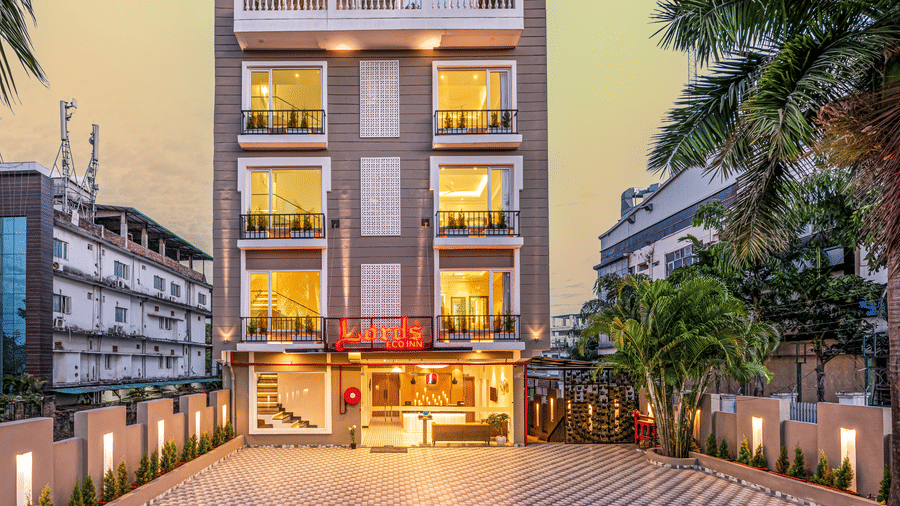 The facade of Lords Eco Inn Dehradun featuring multi storey building with illuminated windows at dusk and a patterned paved driveway flanked by palm trees on both sides at Lords Eco Inn Dehradun.