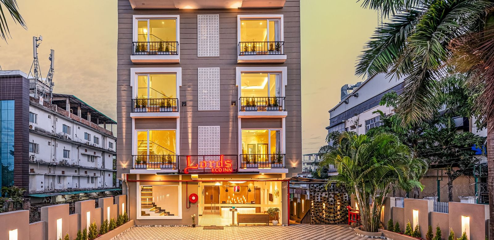 A multi-story building with illuminated windows at dusk, featuring a decorative paved driveway. Palm trees line both sides, with other buildings visible in the background - Lords Eco Inn Dehradun.