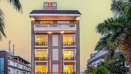 A multi-story building with illuminated windows at dusk, featuring a decorative paved driveway. Palm trees line both sides, with other buildings visible in the background - Lords Eco Inn Dehradun.