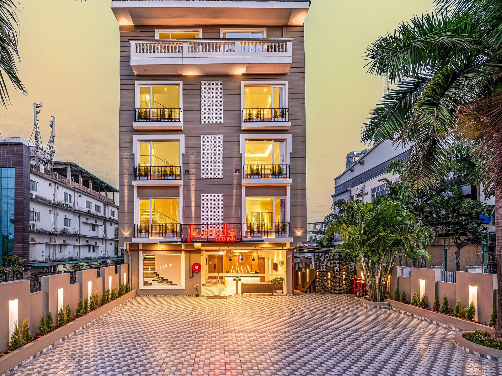 The facade of Lords Eco Inn Dehradun featuring multi storey building with illuminated windows at dusk and a patterned paved driveway flanked by palm trees on both sides at Lords Eco Inn Dehradun.