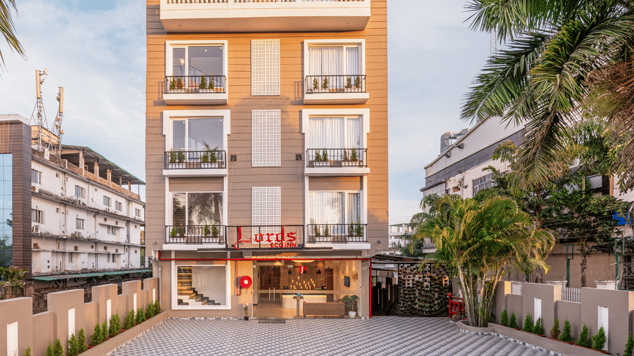 A view of the exterior of Lords Eco Inn Dehradun featuring multi-storey building with a Lords' board sign on top, a paved driveway flanked by palm trees on both its side Lords Eco Inn Dehradun.