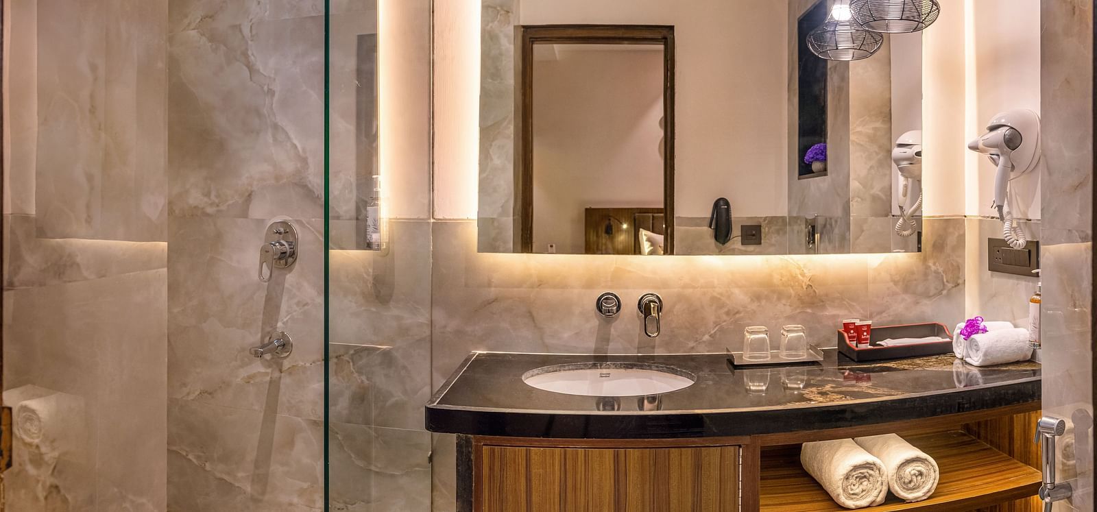 A modern bathroom with a glass-enclosed shower, dark countertop, and a large mirror with lighting. It features a wooden cabinet with open shelves, a white toilet, and hanging pendant lights - Lords Eco Inn Dehradun.