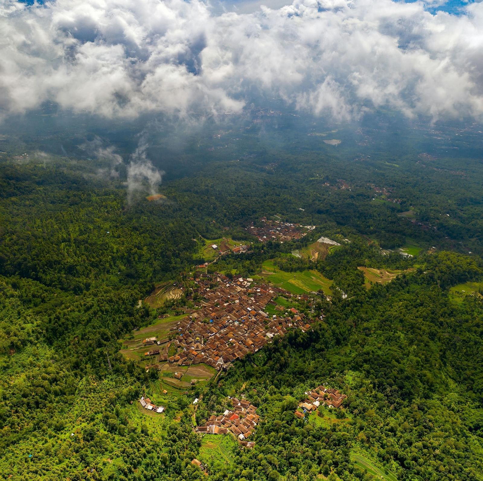 Aerial view of a village surrounded by lush green forests and mountains. White clouds cover the sky.