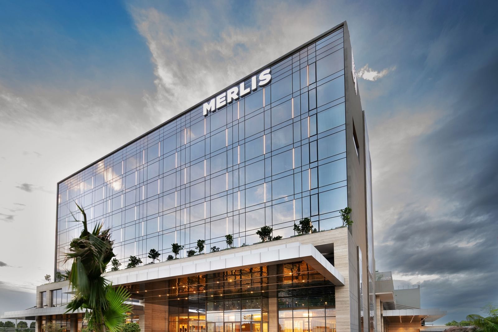 The facade of Merlis Hotel in Coimbatore with with a sleek glass exterior under a vibrant blue sky and a car parked near the entrance