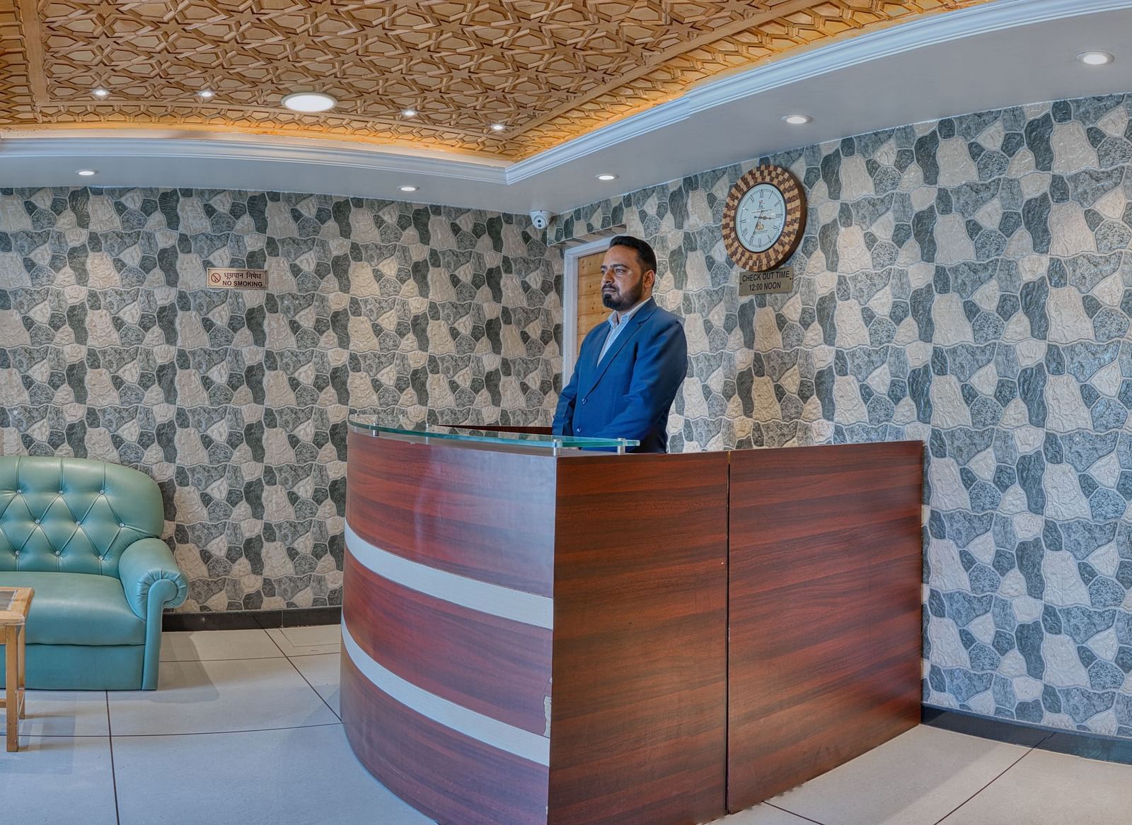 The reception desk at Royal Comfort Regency with a patterned feature wall, green sofa seating and a staff member attending guests.