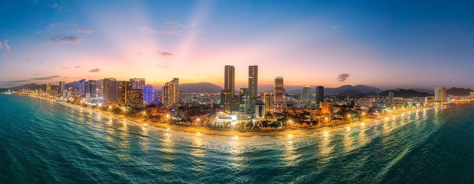 A panoramic cityscape at dusk, featuring a coastline with buildings and lights reflecting on the water.