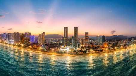 A panoramic cityscape at dusk, featuring a coastline with buildings and lights reflecting on the water.