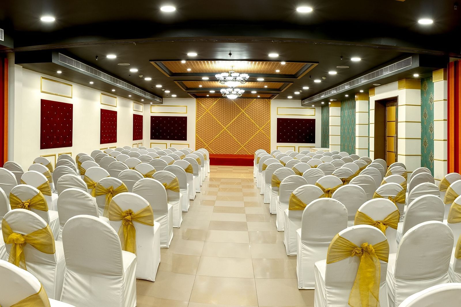 A spacious banquet hall with round tables covered in white cloths and gold chair covers. The hall has a modern design with dark wood accents and a crystal chandelier.