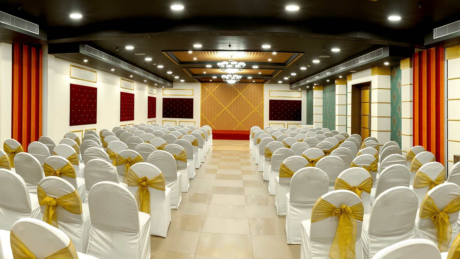 A spacious banquet hall with round tables covered in white cloths and gold chair covers. The hall has a modern design with dark wood accents and a crystal chandelier.