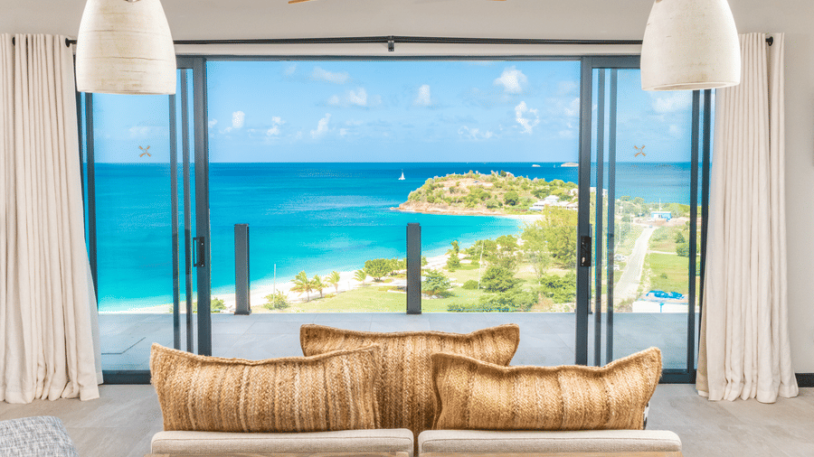 A living area with two sofas, two lounge chairs, a glass sliding door, pendant lights, with a balcony with a view of the sea and the beach, at the Tamarind Hills Resort and Villas, Antigua.