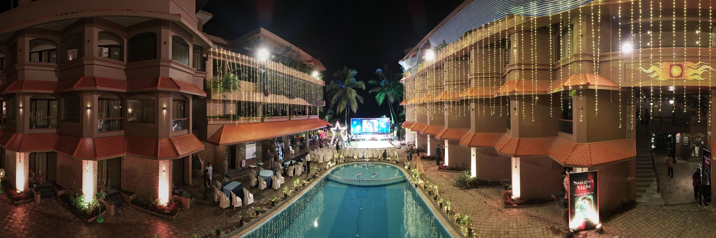 An image of a hotel exterior at night, with vibrant lighting hanging from the building’s roof, illuminating the pool and creating a festive celebration vibe