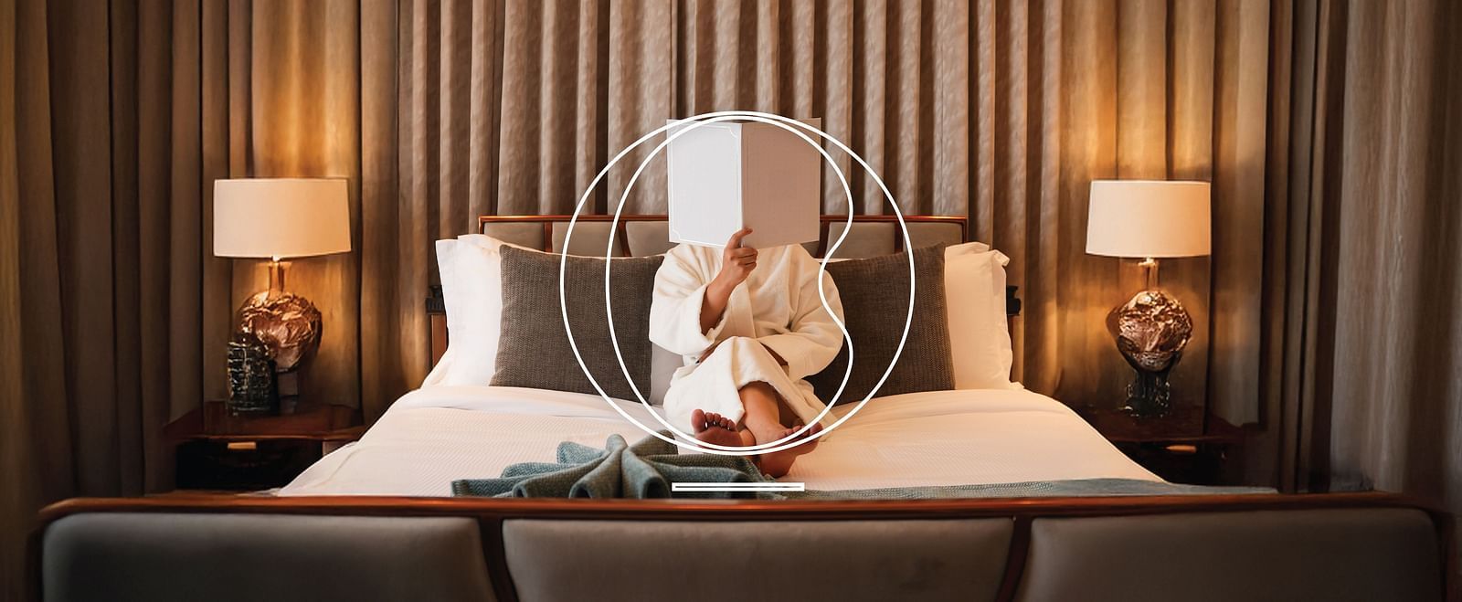 A woman wearing a house coat holding a brochure over her face as she is sitting on the bed of the Suite at Miros Hotels.