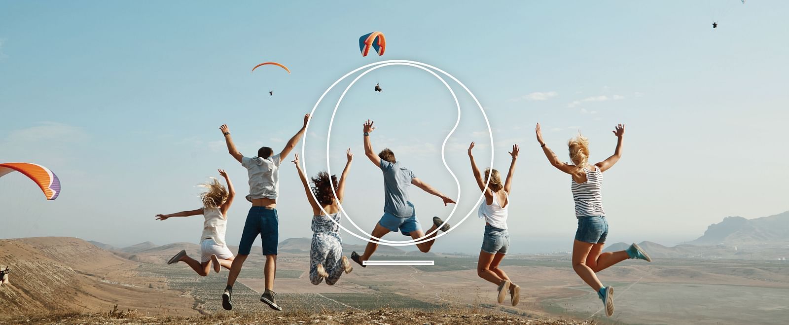 A group of people jumping in joy with several paragliding sails floating in the air.