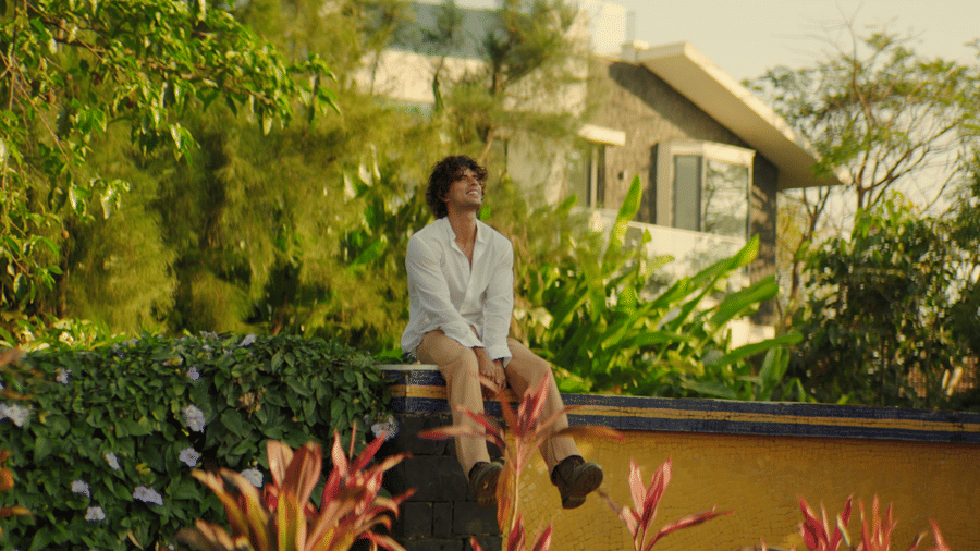 A man in a white shirt sitting peacefully on a wall surrounded by lush green trees and colourful tropical plants under warm sunlight.