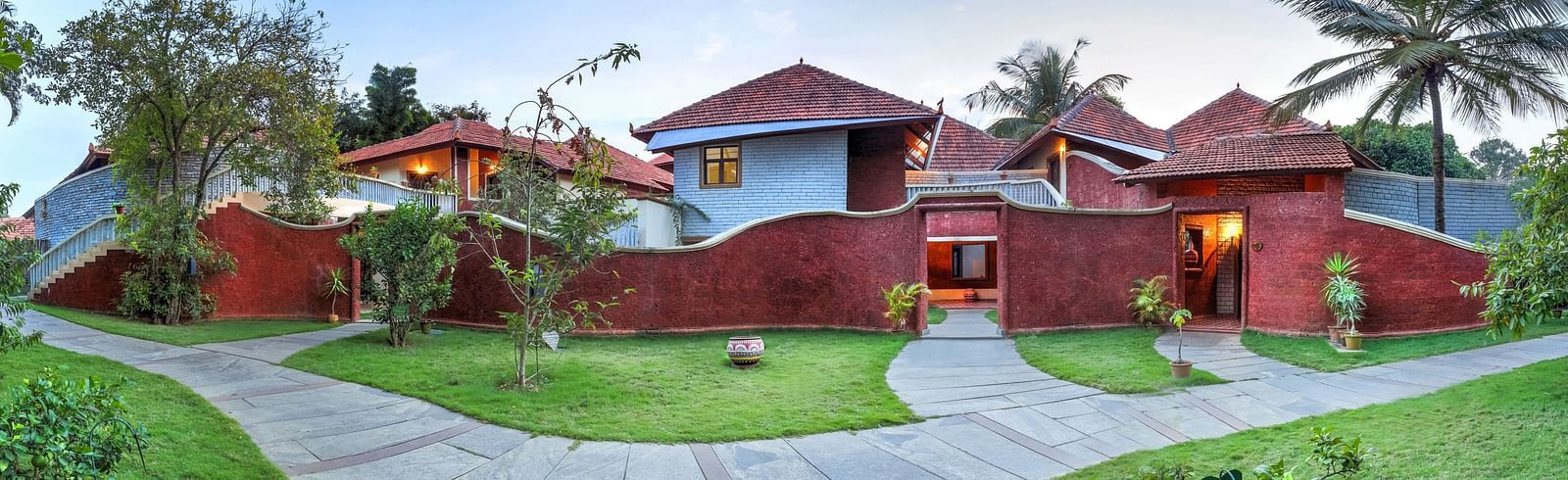 A facade image of a villa which shows thatched huts, palm trees, a pavement, and a lawn - Hoysala Village Resort, Hassan.