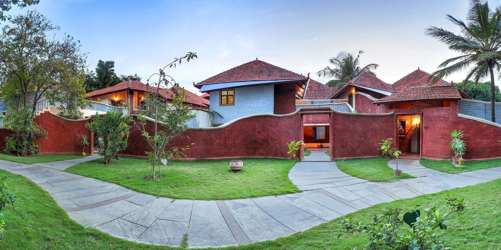 A facade image of a villa which shows thatched huts, palm trees, a pavement, and a lawn - Hoysala Village Resort, Hassan.
