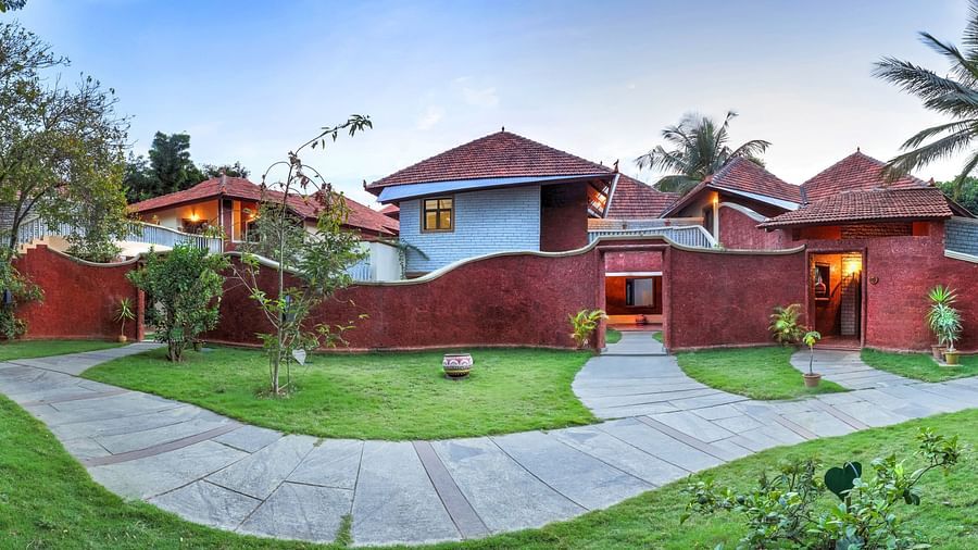 A facade image of a villa which shows thatched huts, palm trees, a pavement, and a lawn - Hoysala Village Resort, Hassan.