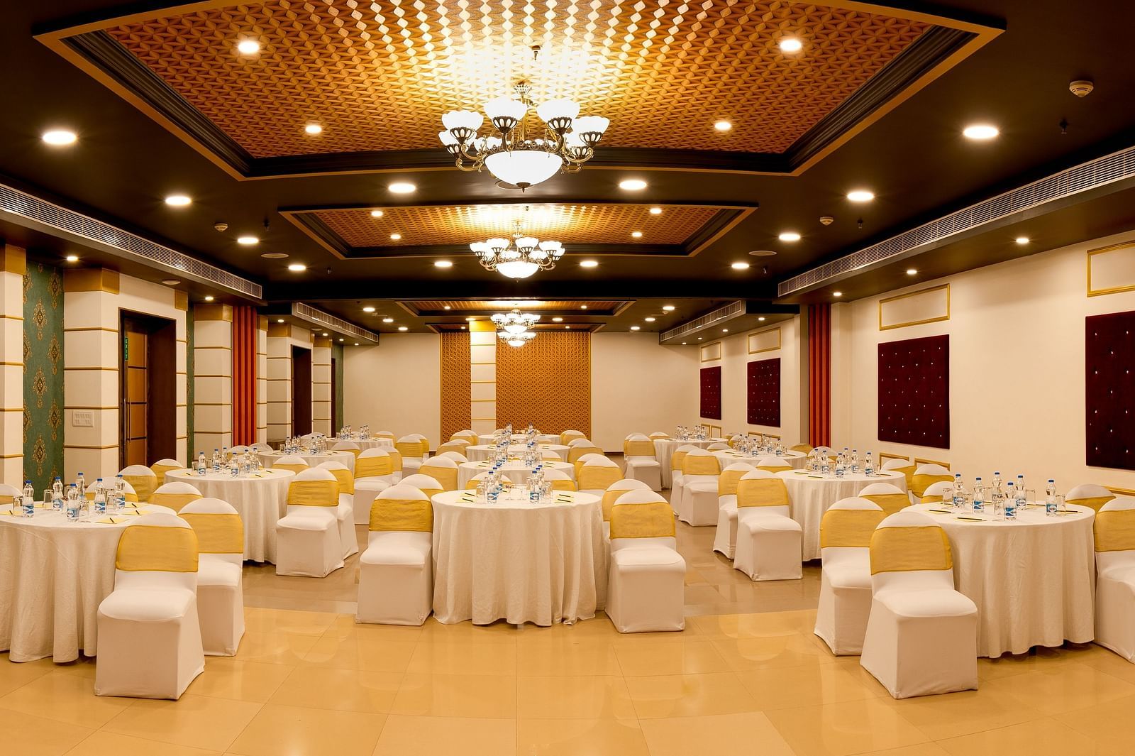 A spacious banquet hall with round tables covered in white cloths and gold chair covers. The hall has a modern design with dark wood accents and a crystal chandelier.