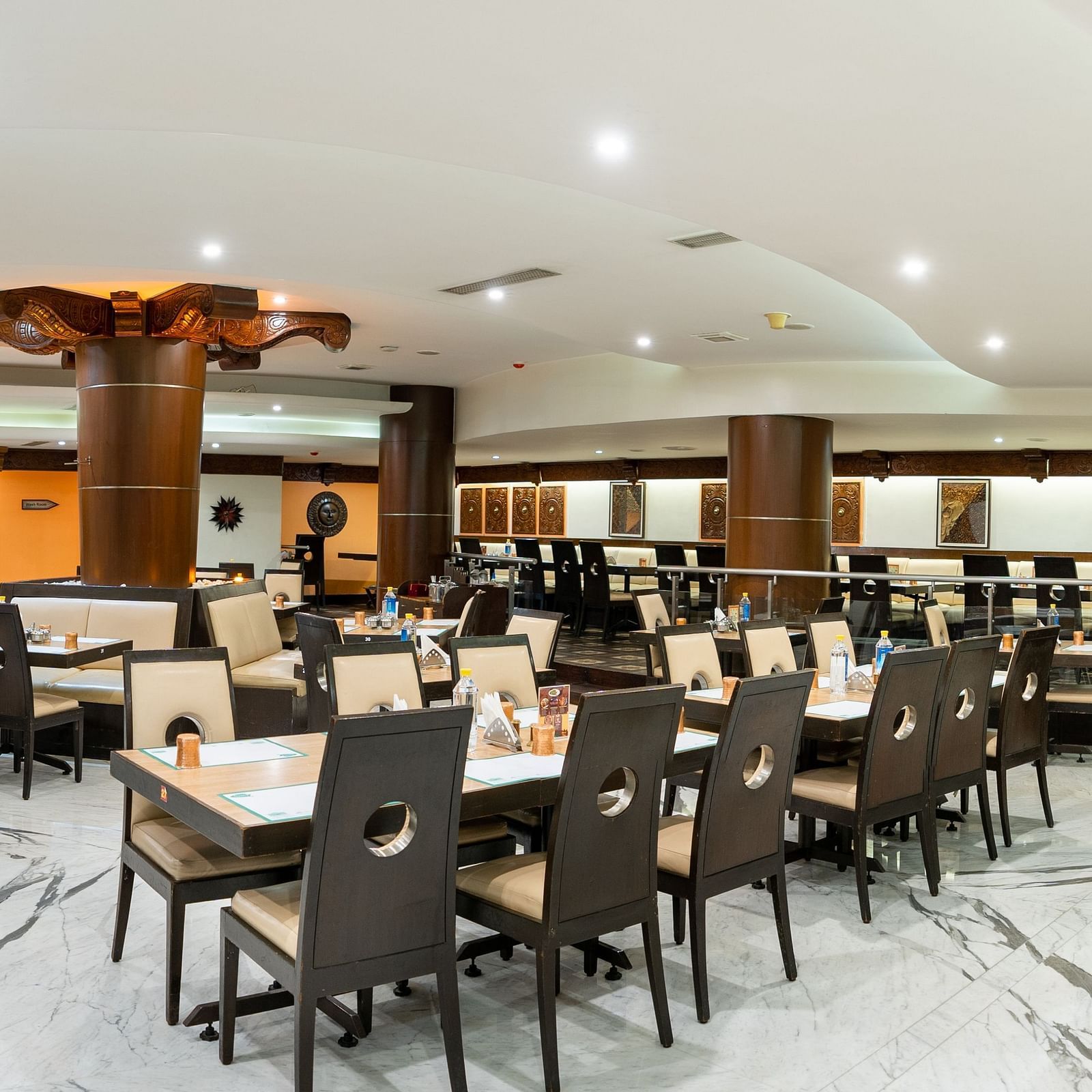 A spacious and well-lit restaurant interior with neatly arranged tables and chairs set for dining under a warm ambient lighting at The President Hotel Jayanagar, Bangalore