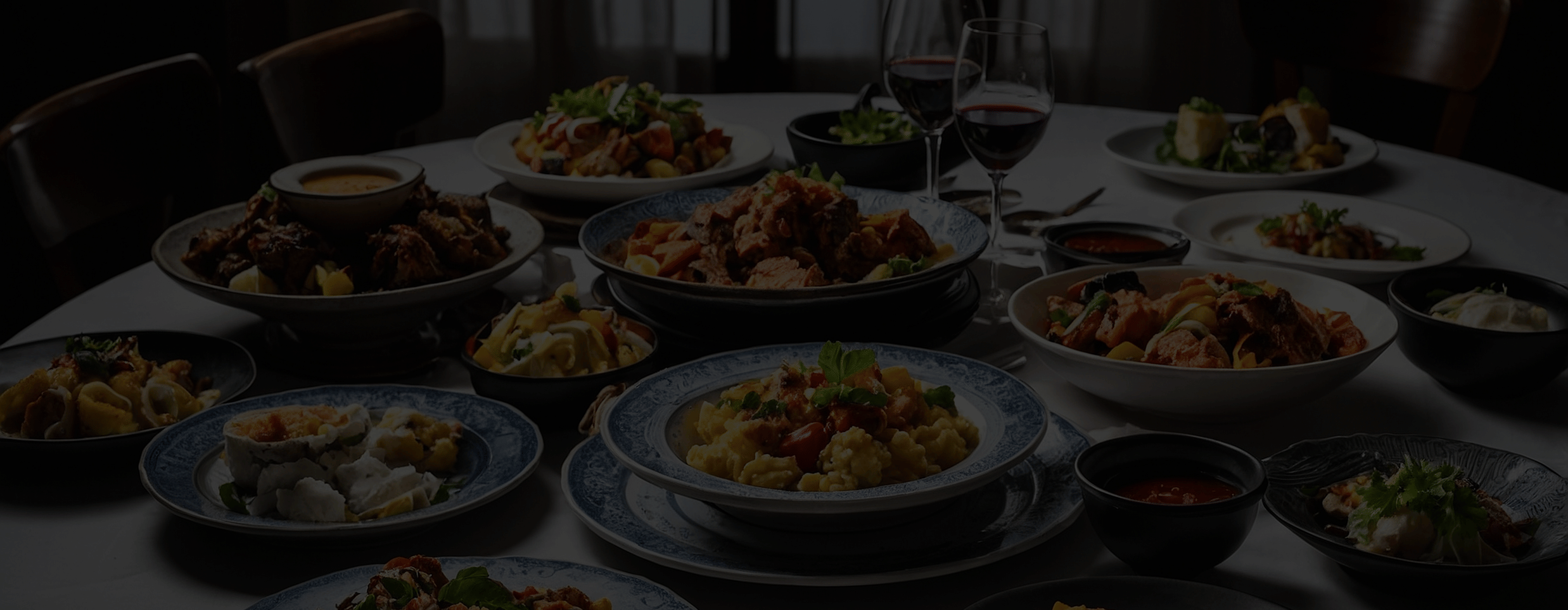Dimly lit dining table set with multiple plates of food, representing a communal banquet-style meal.
