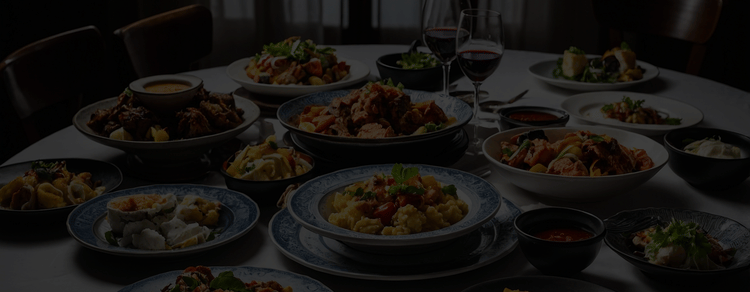 Dimly lit dining table set with multiple plates of food, representing a communal banquet-style meal.