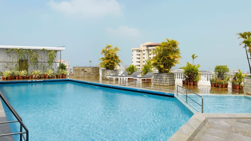 A rooftop swimming pool with clear blue water, surrounded by potted plants and city views under a bright sky - Abad Plaza, MG Road, Shenoy Kochi