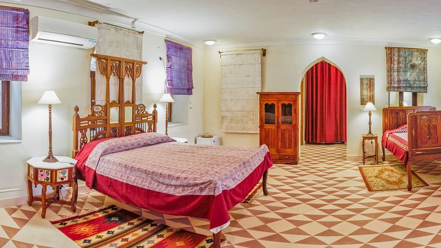 Spacious bedroom with two beds, wooden furniture, patterned flooring, windows with blinds, and a doorway with a curtain at Tijara Fort-Palace - 19th Century, Alwar.