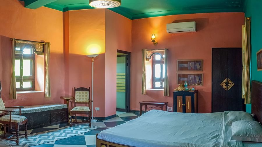Bedroom with a bed, wooden furniture including chairs and a cabinet, windows, and an air conditioning unit at Tijara Fort-Palace - 19th Century, Alwar.