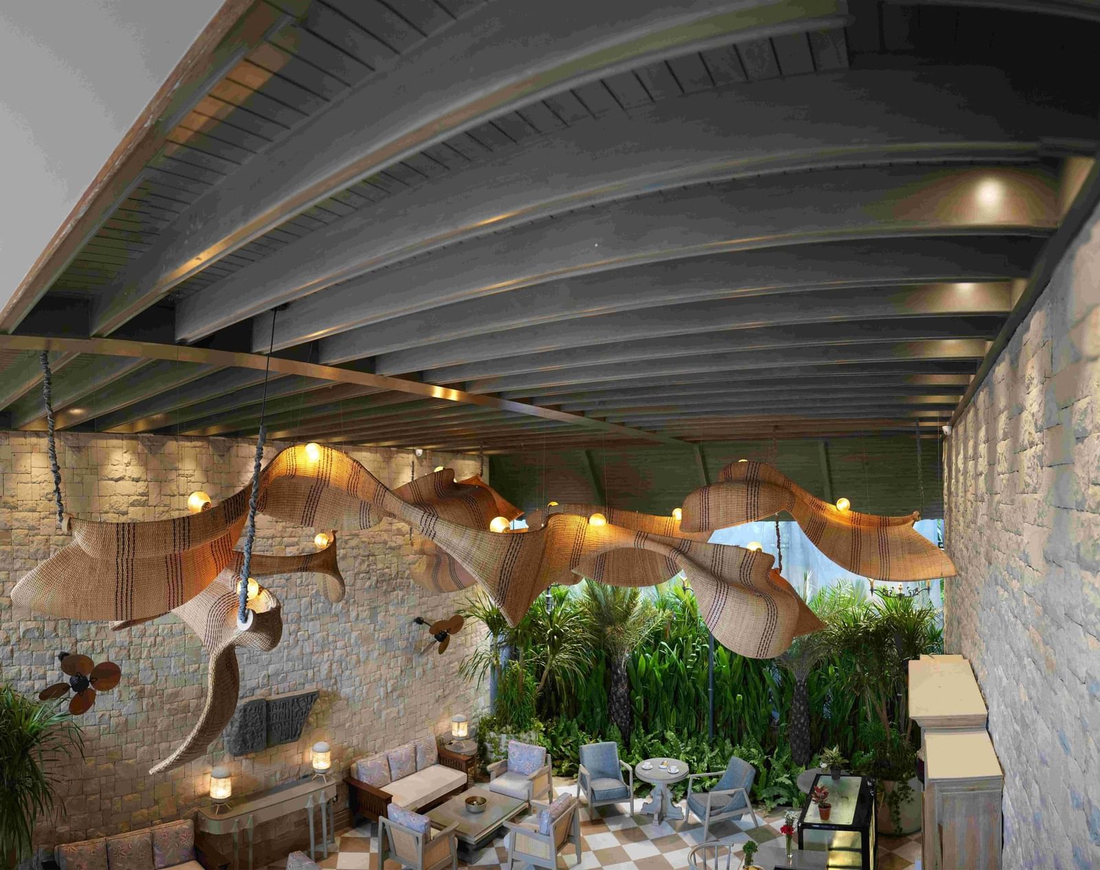 An expansive, high-angle view of the charming Suenos De Cafe at The Evren, Vagator. The space features a checkered floor, comfortable seating, and a striking, large, woven light fixture. The walls are made of stone, and a lush green garden is visible in th