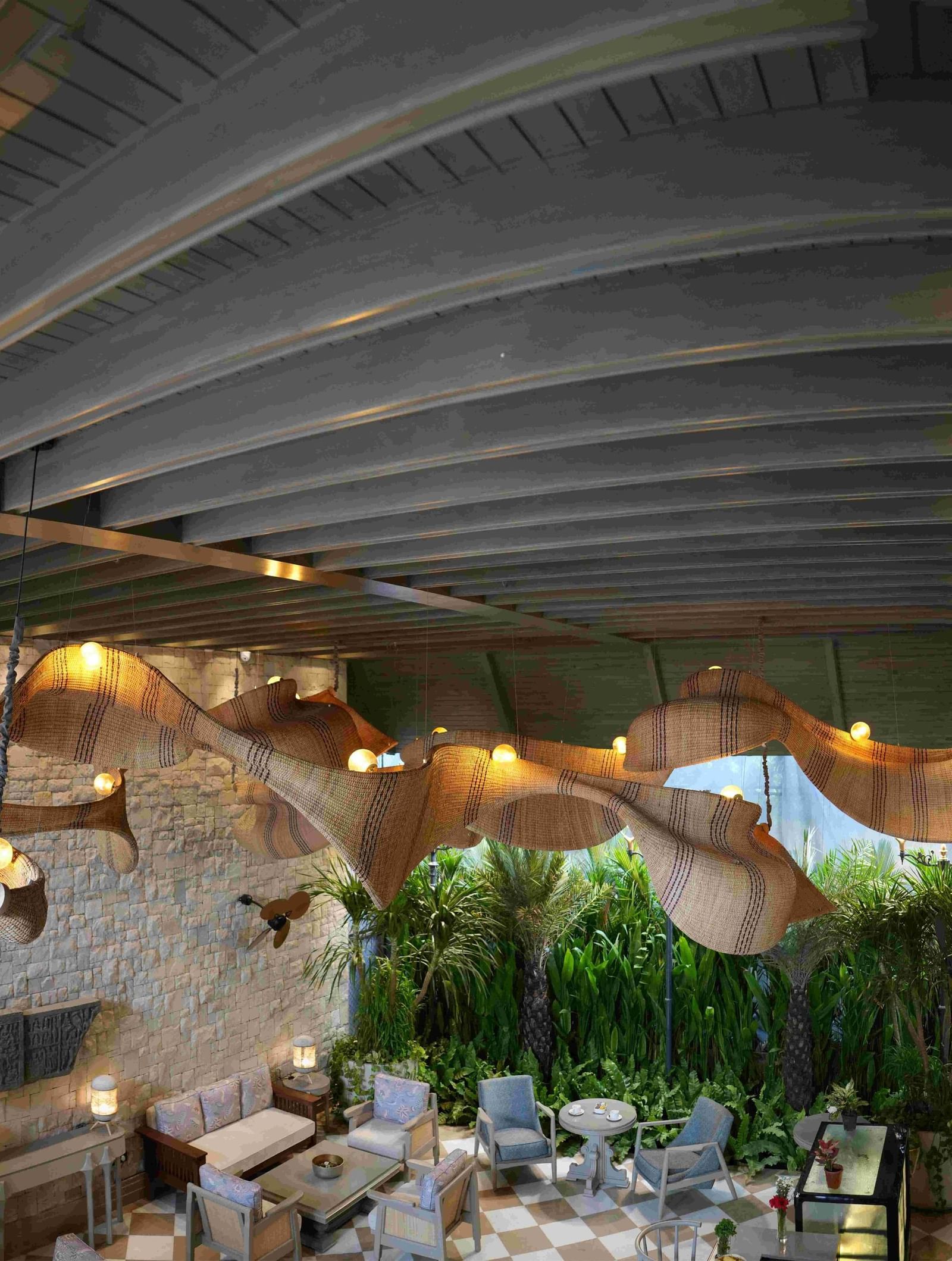 An expansive, high-angle view of the charming Suenos De Cafe at The Evren, Vagator. The space features a checkered floor, comfortable seating, and a striking, large, woven light fixture. The walls are made of stone, and a lush green garden is visible in th