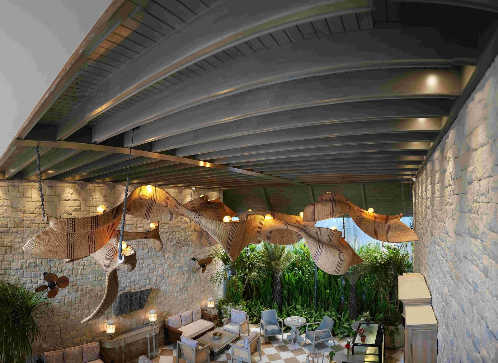 An expansive, high-angle view of the charming Suenos De Cafe at The Evren, Vagator. The space features a checkered floor, comfortable seating, and a striking, large, woven light fixture. The walls are made of stone, and a lush green garden is visible in th