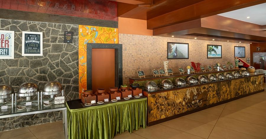 An elaborate buffet setup featuring stainless steel serving dishes, traditional clay pots at the non-vegetarian counter, and decorative wall art, with signs promoting cold beer and a quirky tequila quote at Uday Samudra Leisure Beach Hote
