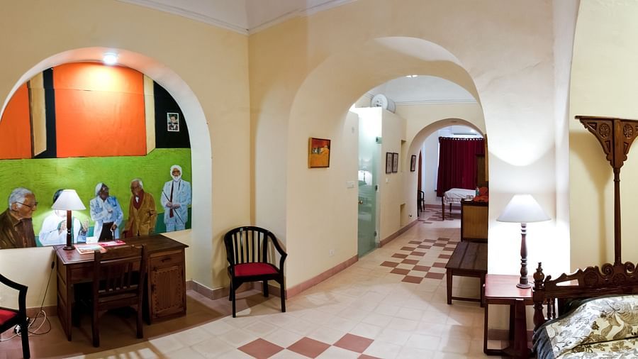 A hallway with arched openings, a desk with chairs and artwork, and a bed with a lamp at Tijara Fort-Palace, Alwar.