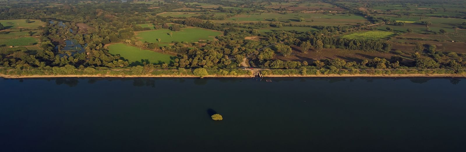 An aerial view of a dark body of water with a small object floating in it, bordered by a forested landscape- Shahpura Bagh.