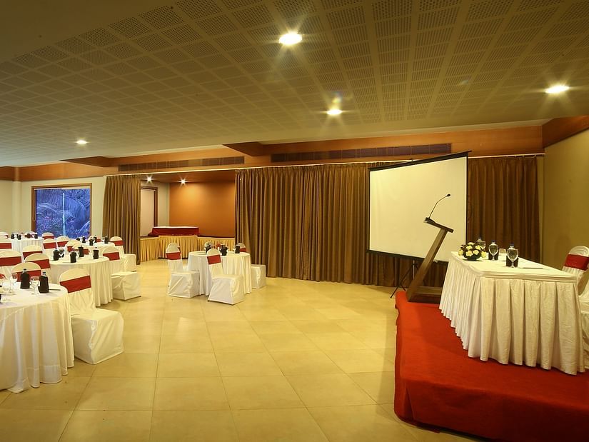 Marigold, the event hall with neatly arranged chairs, a stage with a table and chairs for guests, a microphone stand, and a projector screen, highlighted by glass windows and warm lighting at Uday Suites, Trivandrum
