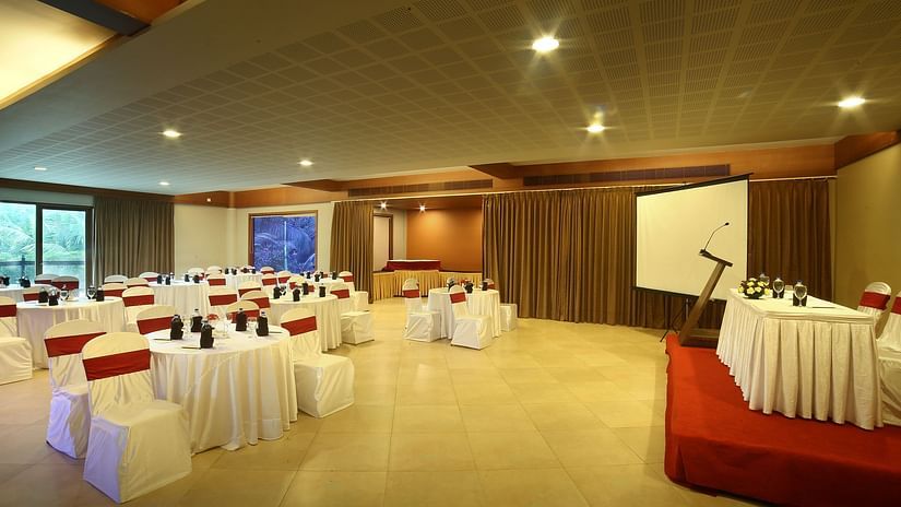 An image of an event hall with neatly arranged chairs, a stage with a table and chairs for guests, a microphone stand, and a projector screen, highlighted by glass windows and warm lighting - Uday Suites, Trivandrum
