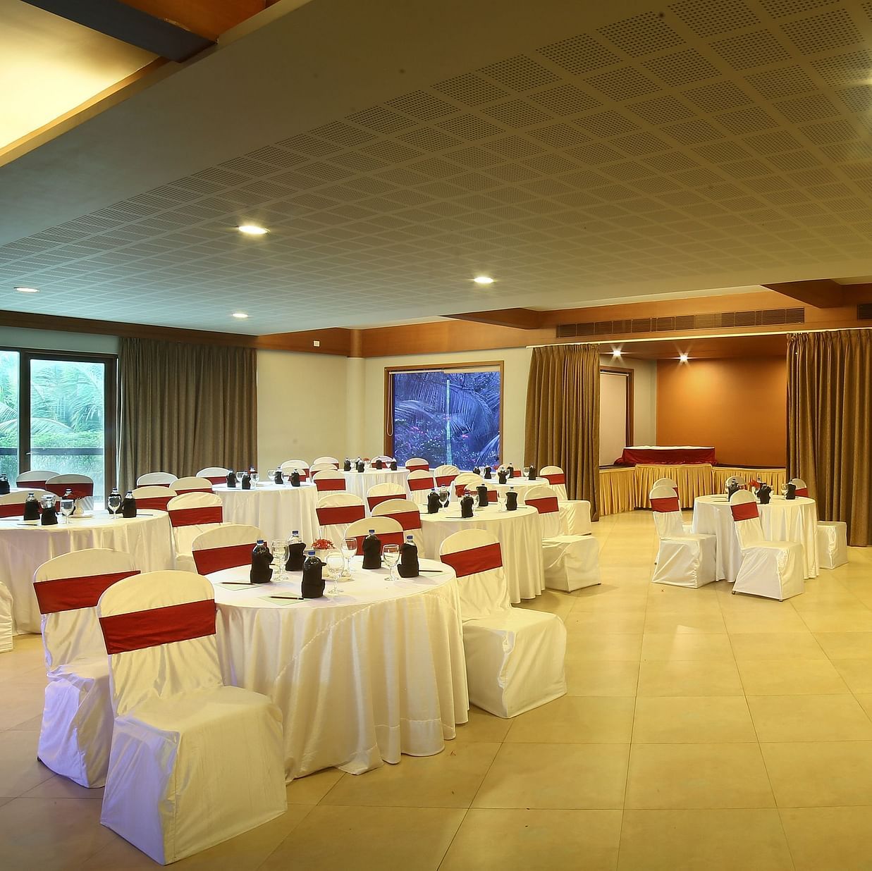 Marigold, the event hall with neatly arranged chairs, a stage with a table and chairs for guests, a microphone stand, and a projector screen, highlighted by glass windows and warm lighting at Uday Suites, Trivandrum