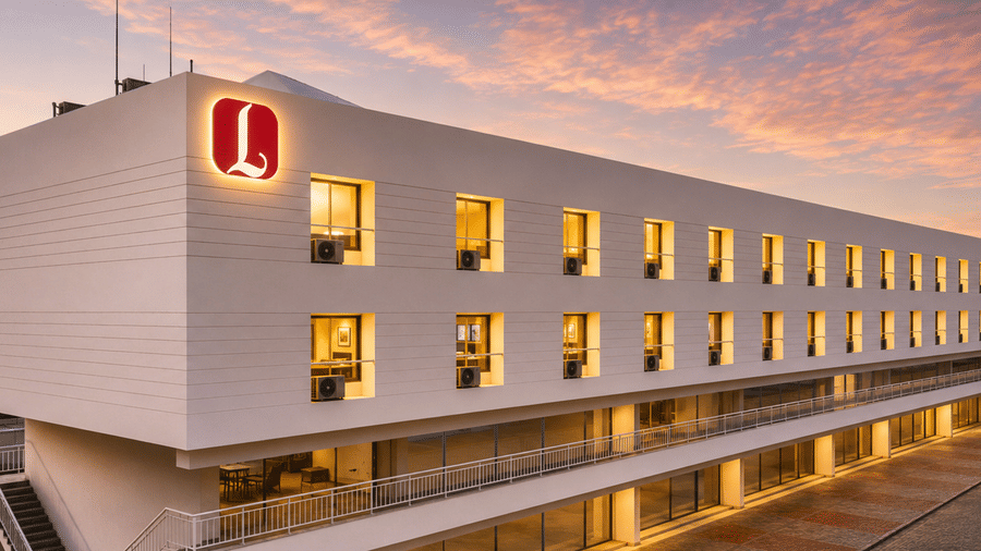 Exterior view of Angel Lords Eco Inn Amreli at dusk, showcasing a long modern white building with warm illuminated windows, a red rooftop logo, and a sunset sky in the background.
