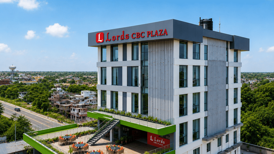 Elevated exterior view of Lords CBC Plaza, Chitwan showing a contemporary multi-story white and grey hotel building with an open terrace, outdoor seating, and surrounding greenery along a main road.