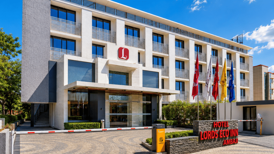 Exterior daytime view of the Hotel Lords Eco Inn Ambaji featuring a paved entrance driveway, security barrier, modern white facade, and colourful flagpoles under a clear blue sky.