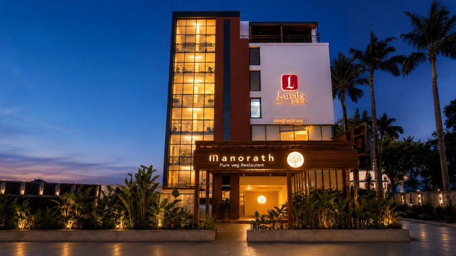 Exterior dusk view of Lords Eco Inn Belagavi featuring an illuminated multi-story building facade, glass-walled stairwell, palm trees, and warm entrance lighting under a deep blue twilight sky.