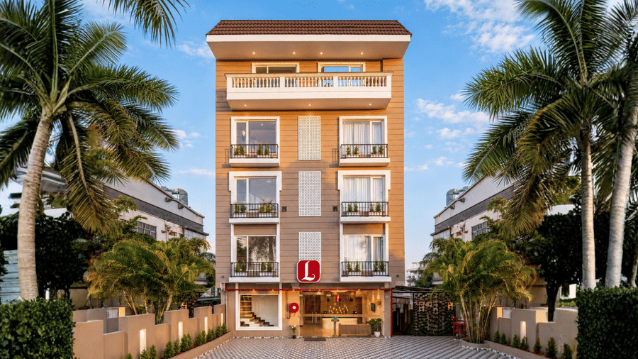 Exterior view of Lords Eco Inn Dehradun featuring a multi-story hotel building with balconies, paved entrance, and surrounding palm trees under a clear sky.