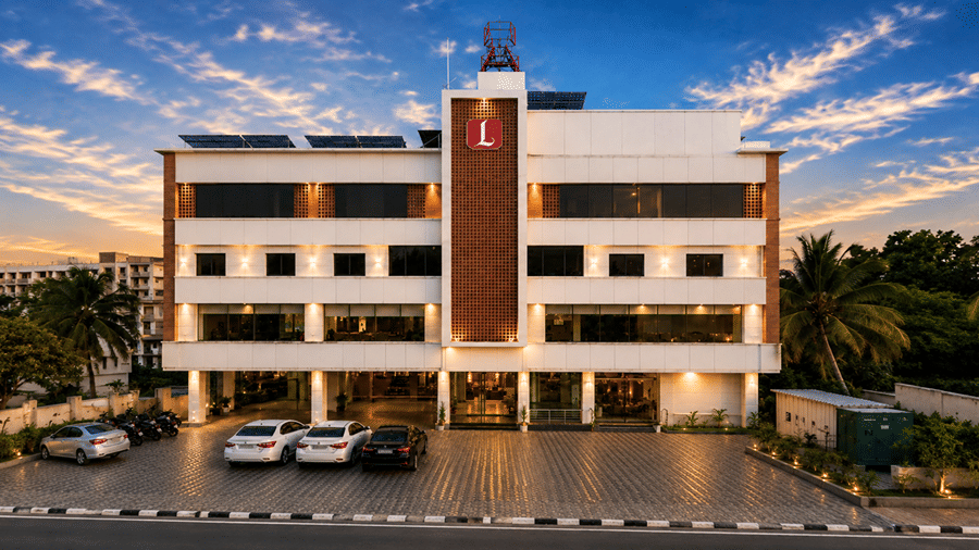 Exterior view of Lords Eco Inn Navsari showing a illuminated multi-story building at dusk, featuring a red logo, ground-floor entrance, parked cars, and surrounding palm trees against a dramatic sky.