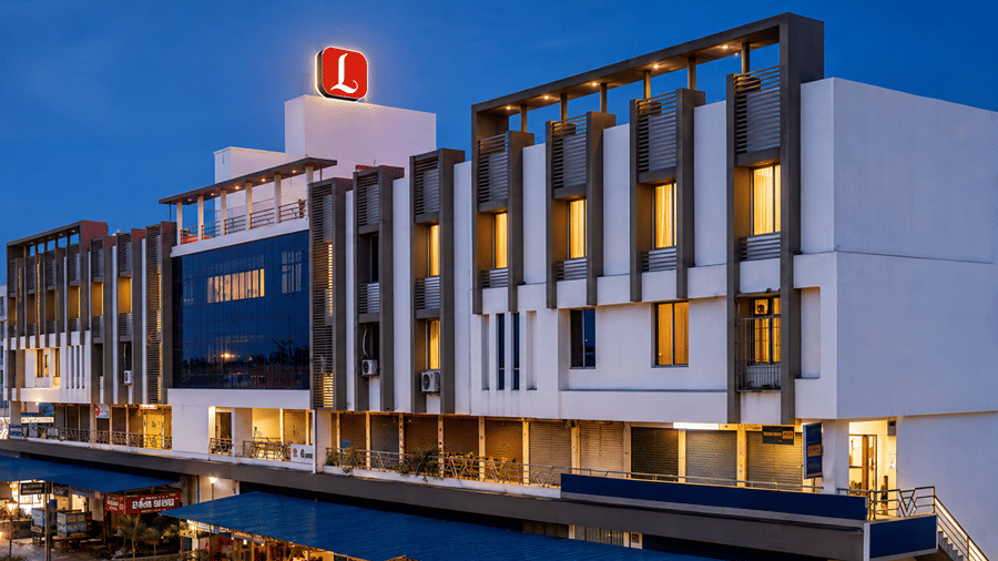 Illuminated exterior view of Lords Eco Inn Shapar Rajkot at night featuring a long multi-story facade with vertical architectural fins, warm lighting from windows, and a illuminated red rooftop logo.