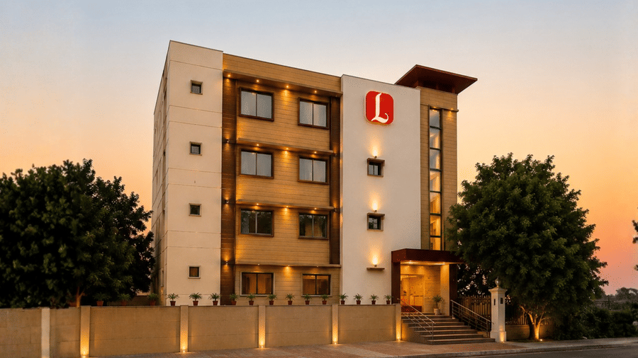 Exterior sunset view of Lords Inn Amritsar showing a brightly lit multi-story hotel building with a red logo and lush green trees on either side under a warm twilight sky.
