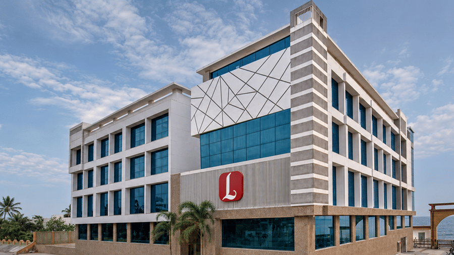 Exterior view of Lords Inn Porbandar featuring a modern multi-story hotel building with glass windows, textured facade panels, a red brand logo, and a clear blue sky.