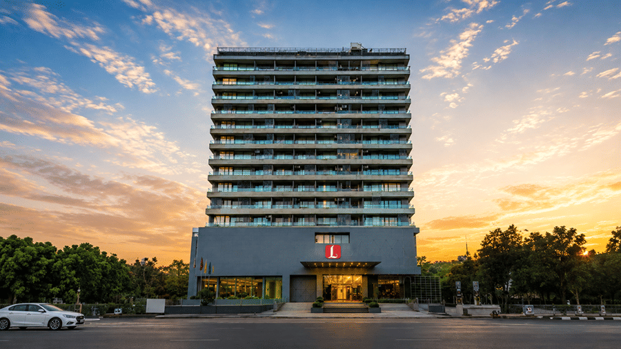 Exterior view of the Lords Inn Rajkot hotel at sunset, showing the tall, modern building facade with numerous well-lit windows against a gradient orange and blue sky, with trees in the foreground.