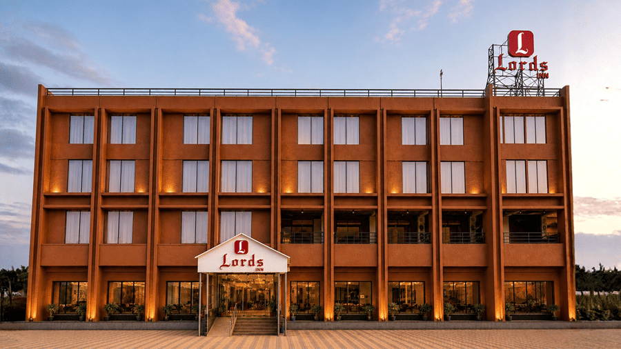 Exterior front view of Lords Inn Sanand Gujarat showing a multi-story brown building with warm facade lighting, entrance canopy, and rooftop branding at dusk.