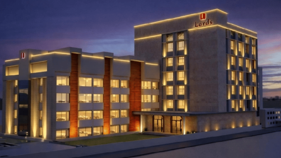 Exterior view of Lords Inn Somnath at twilight featuring a multi-story modern building with glowing yellow facade accent lighting, illuminated windows, and rooftop logo set against a purple-tinged sky.